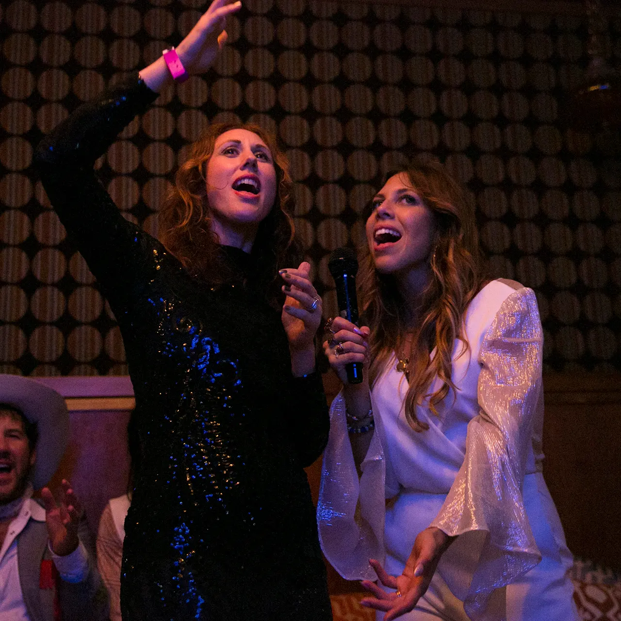 Two people singing karaoke with expressive gestures in warm-lit venue, one in sequins, one in flowing white