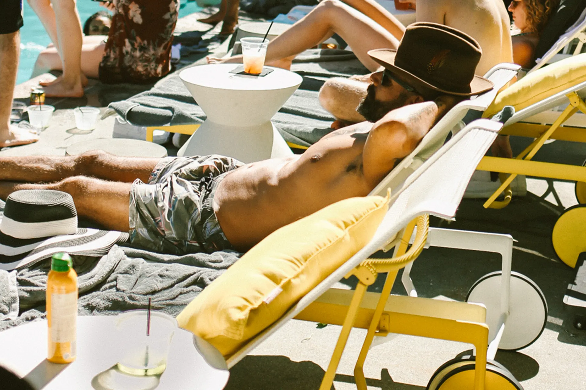 Person lounging shirtless in wide-brimmed hat by poolside with patterned trunks, drinks, and summer accessories