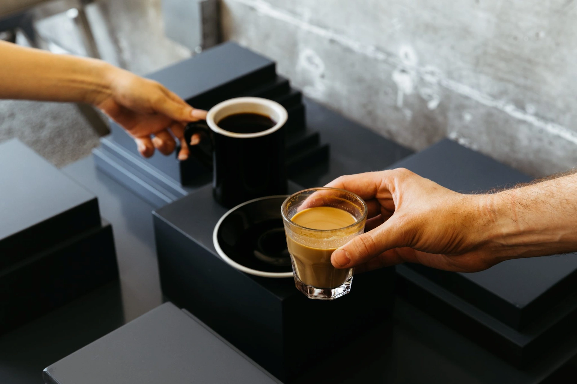 Two hands holding black coffee and latte in minimalist café with concrete wall and black platforms