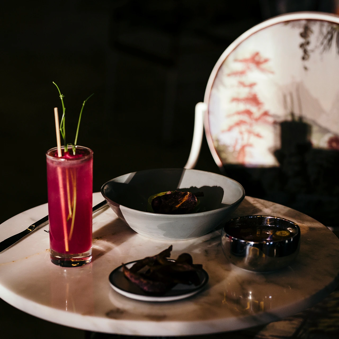 Elegant marble table with pink cocktail, grilled dish, and dramatic shadows in dimly lit setting