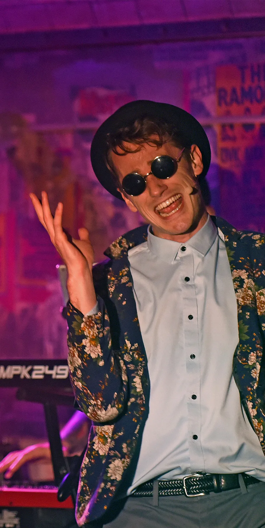 Performer in floral jacket and sunglasses singing near keyboard in colorful, poster-filled music space