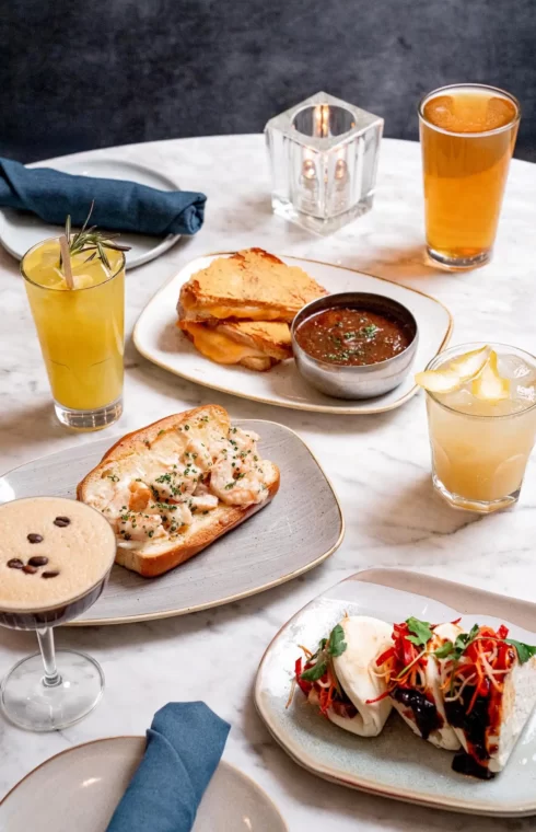 Marble table with assorted dishes, bao buns, sandwiches, cocktails, and beer arranged for a shared meal.