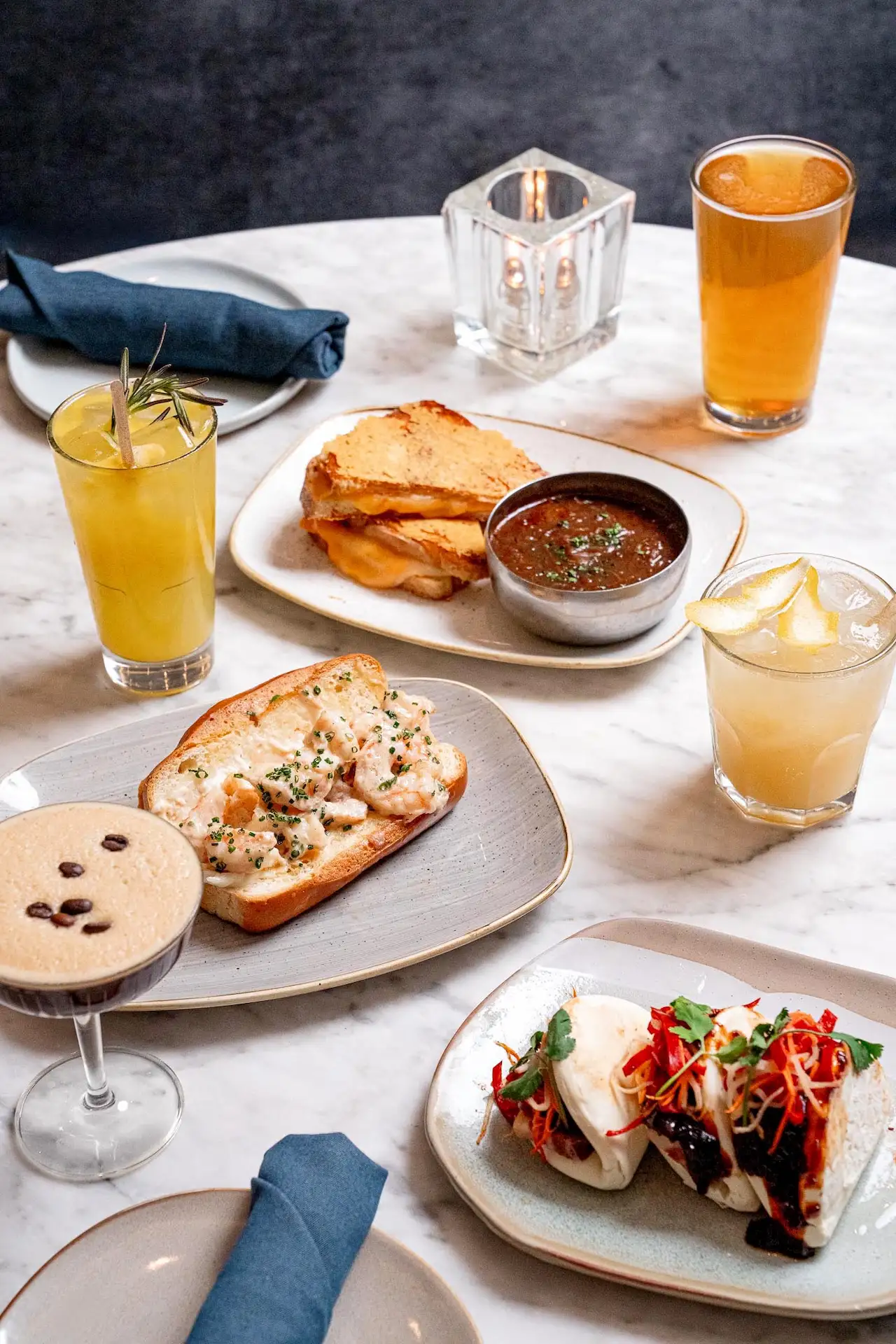 Marble table with assorted dishes, bao buns, sandwiches, cocktails, and beer arranged for a shared meal.