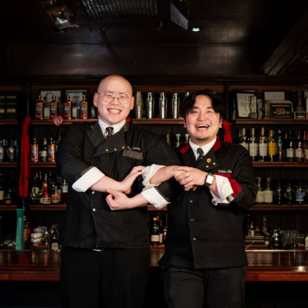 Two smiling bartenders pose behind a bar, linking arms in matching black uniforms with liquor bottles behind them.
