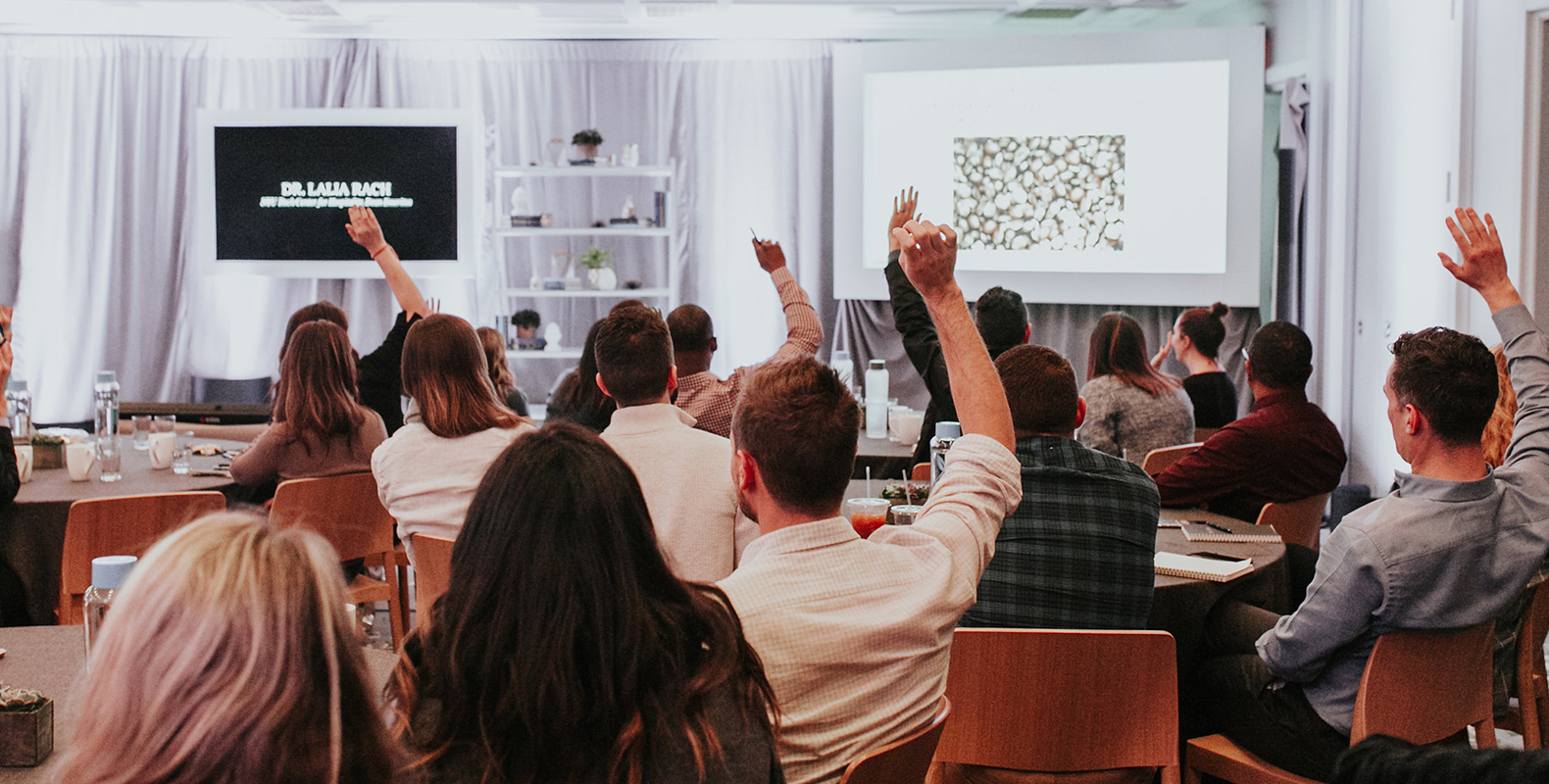 picture of people raising hands while sitting on a brow color chair and table with two big screens