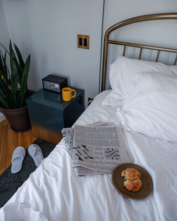 A pastry and folded newspaper sit on a hotel bed with slippers on the floor and a coffee cup on the side table