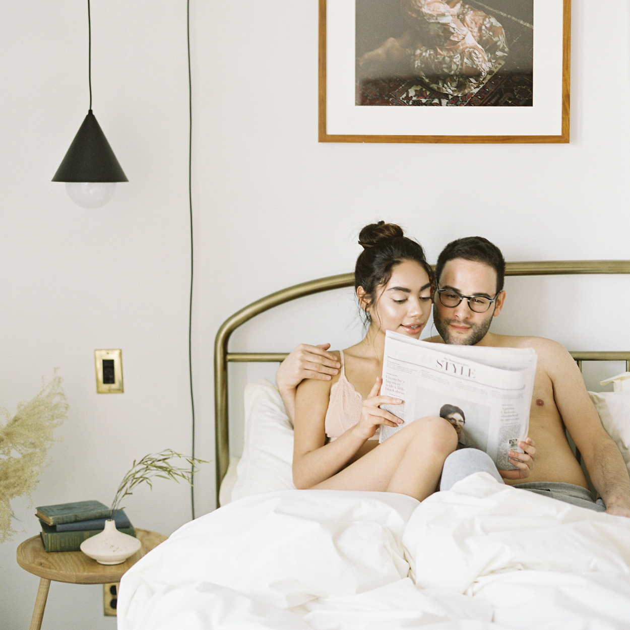Two people sitting up in bed reading a newspaper together