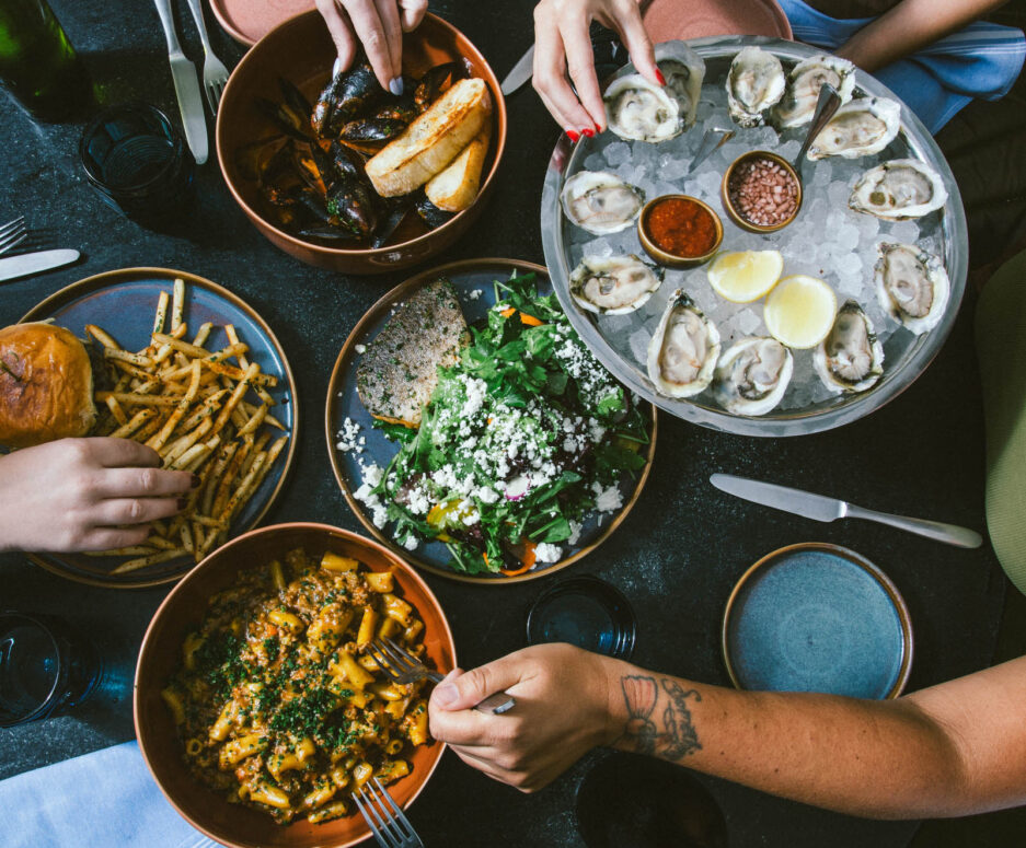 A plate of burger with fries, a cold dish with a dozen oysters on ice, a pasta dish, a salad dish, and a bowl of mussels