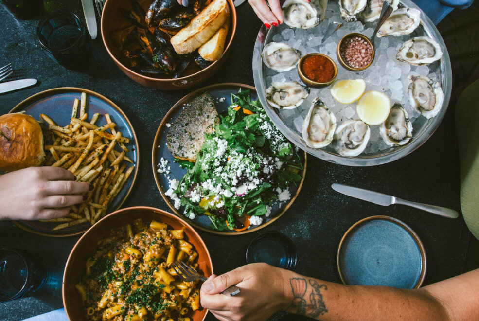 A plate of burger with fries, a cold dish with a dozen oysters on ice, a pasta dish, a salad dish, and a bowl of mussels