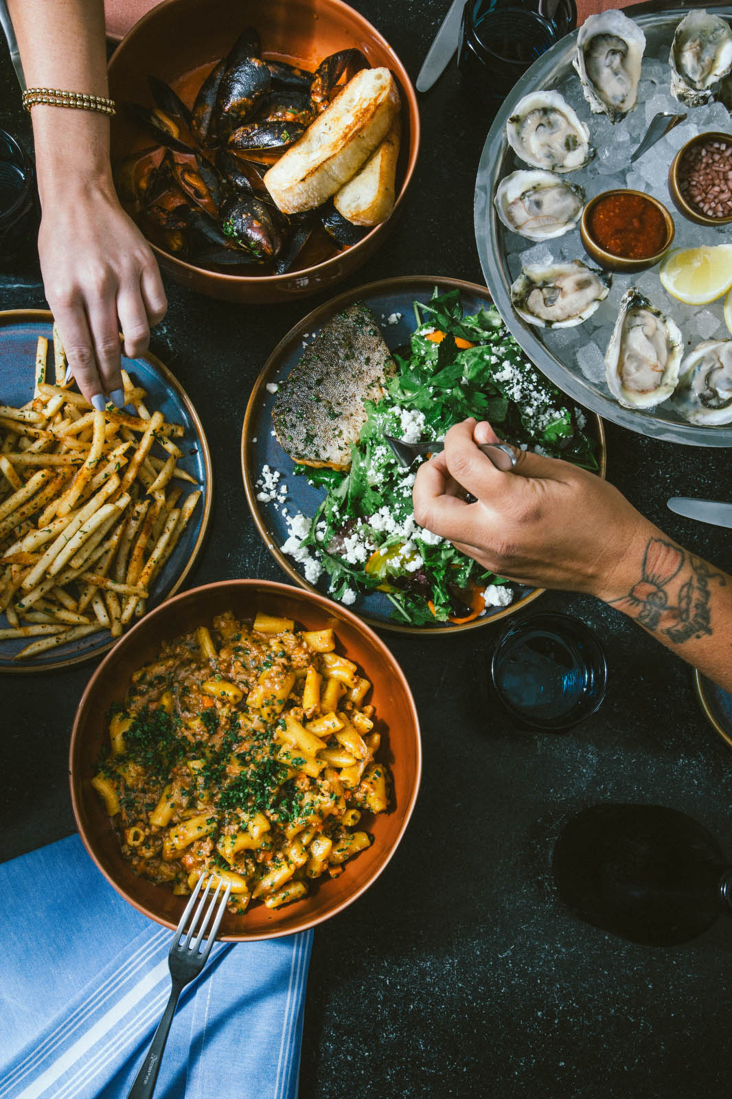 A person's hand reaching out for french fries, and another hand with a fork reaching for salad, next to a cold dish with a dozen oysters on ice, a pasta dish, and a bowl of mussels