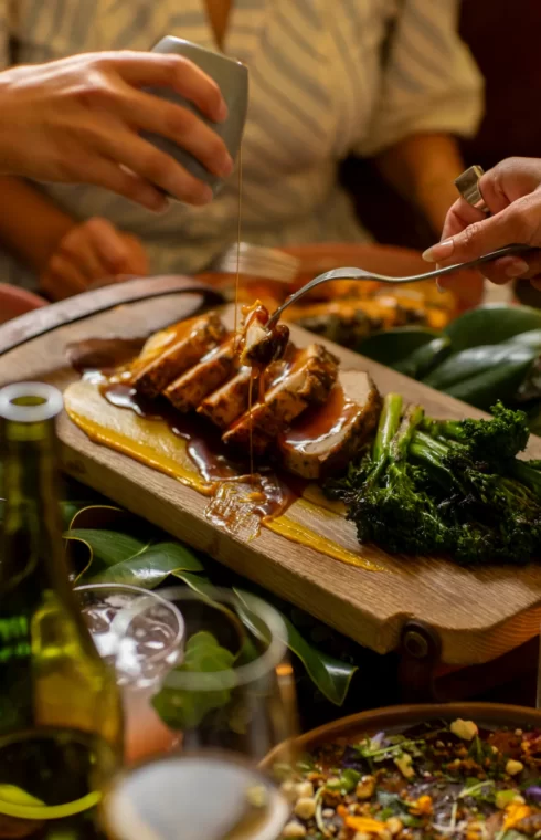 Close-up of sliced meat with sauce, broccolini, and puree on a wooden board at a festive shared meal.