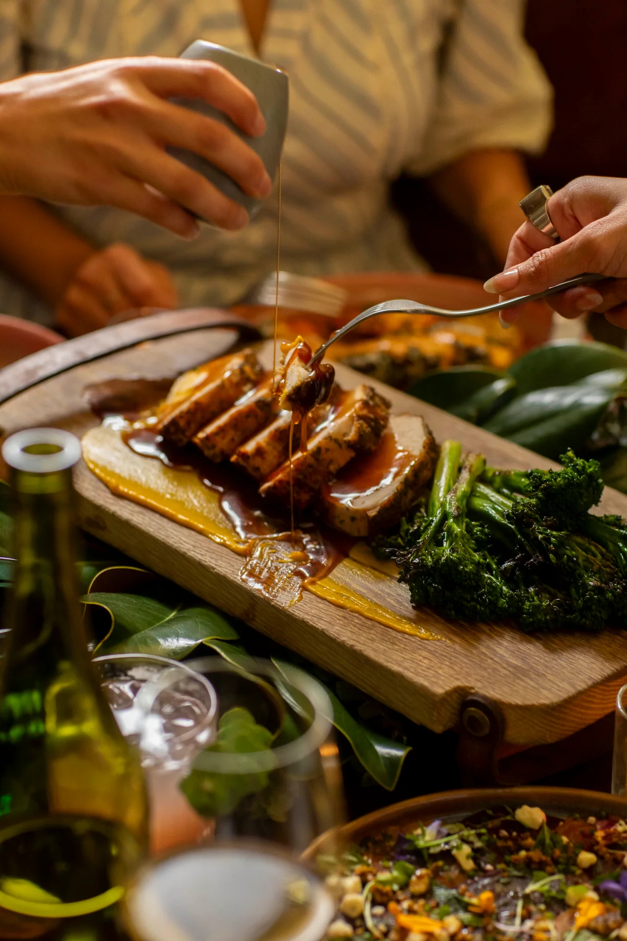 Close-up of sliced meat with sauce, broccolini, and puree on a wooden board at a festive shared meal.