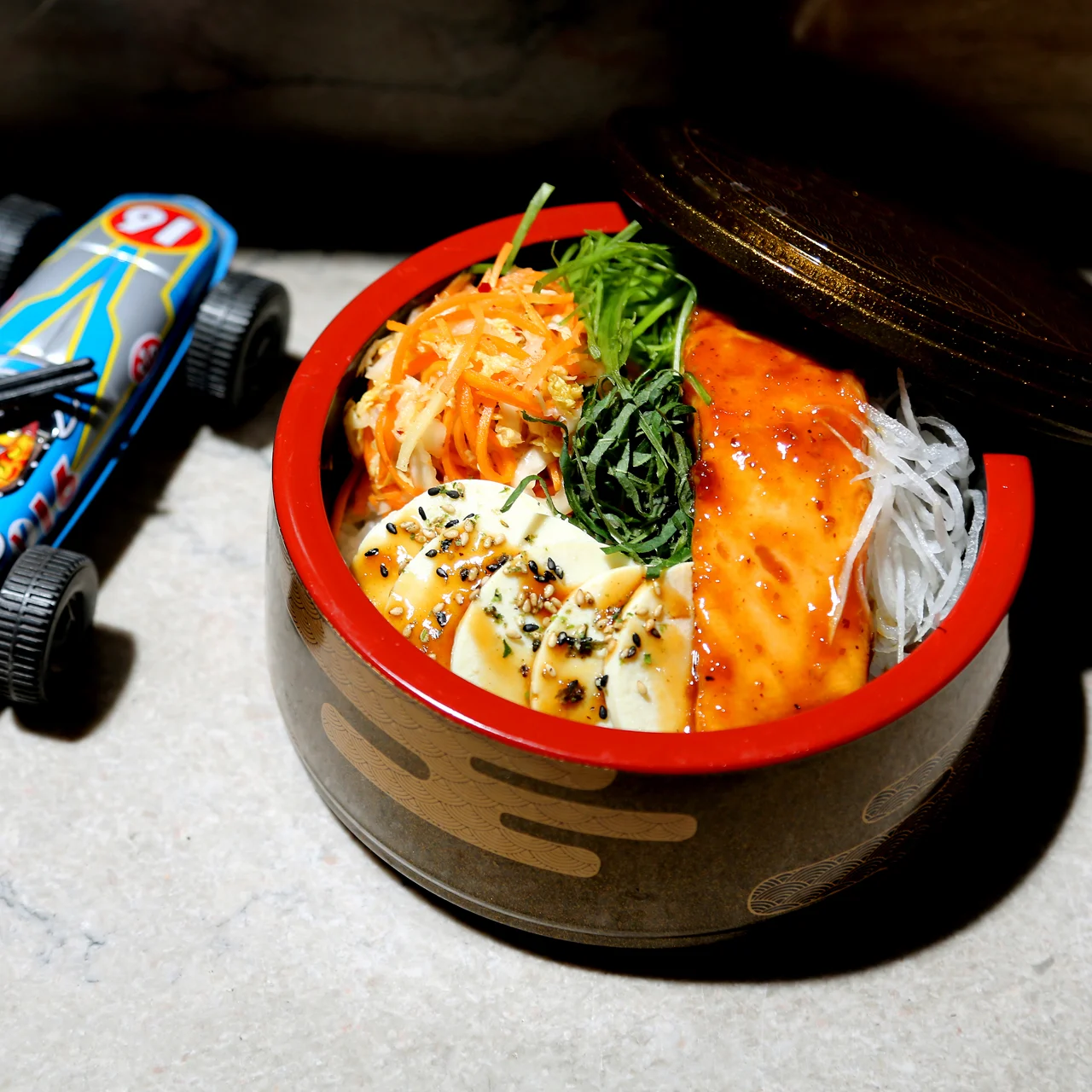 Korean-style bowl with colorful toppings beside toy race car on table, blending food and playfulness