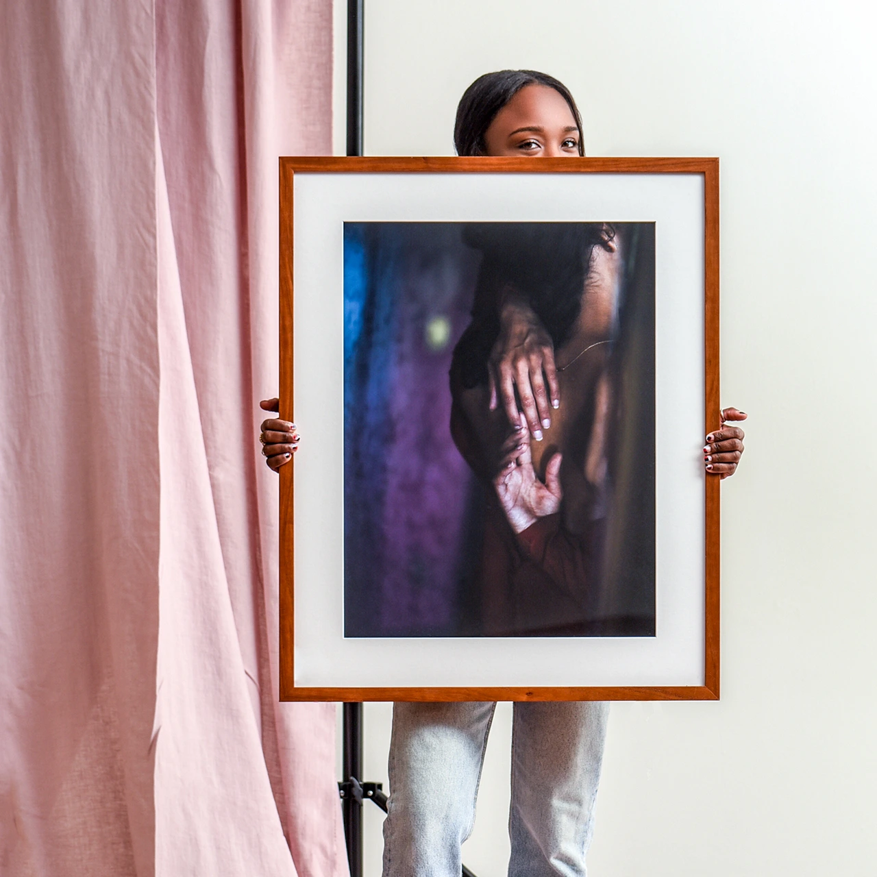 Person holding framed photo of hands in intimate gesture, standing against pink curtain and pale wall