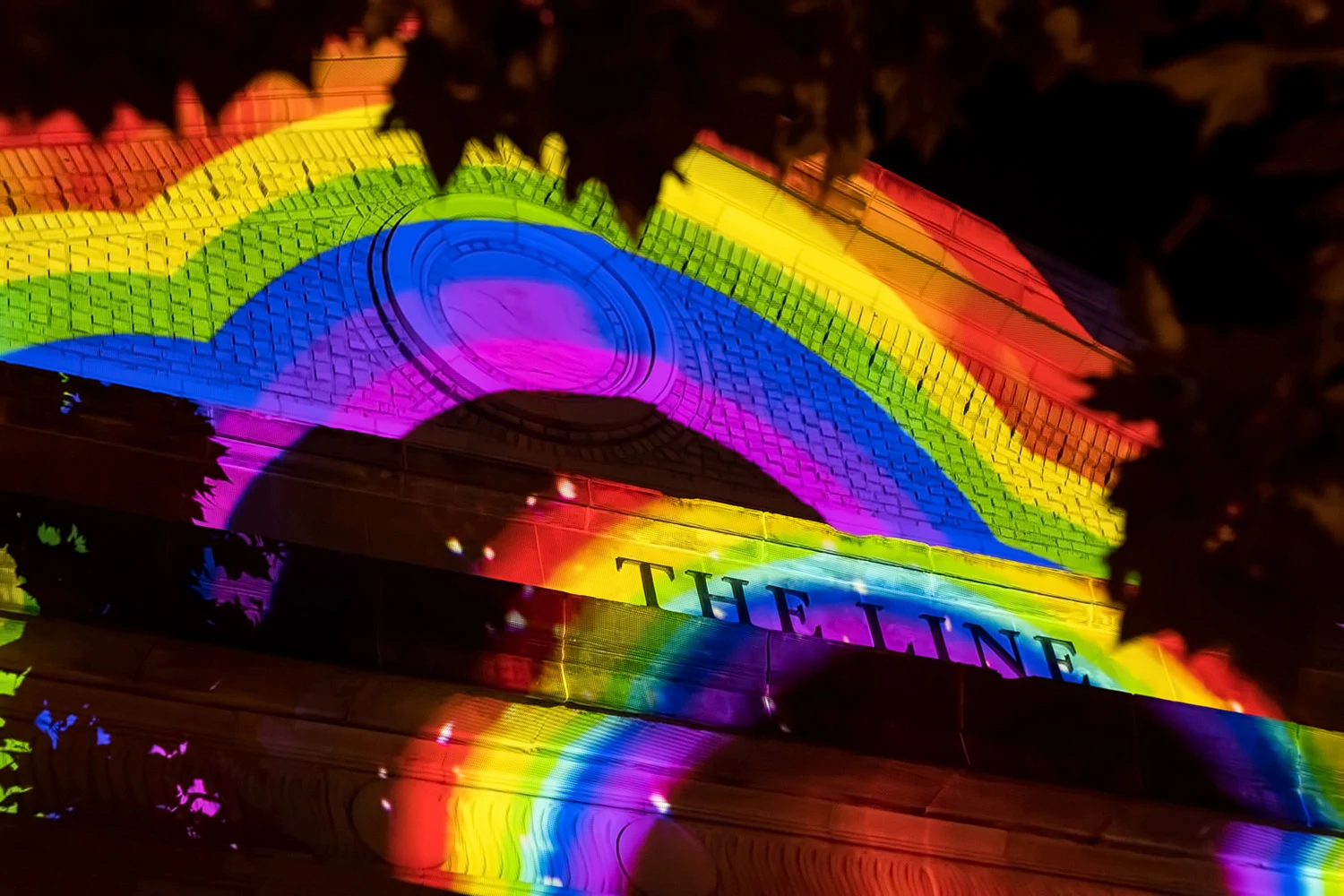Building facade lit with rainbow light projections and THE LINE signage, framed by silhouetted leaves at night