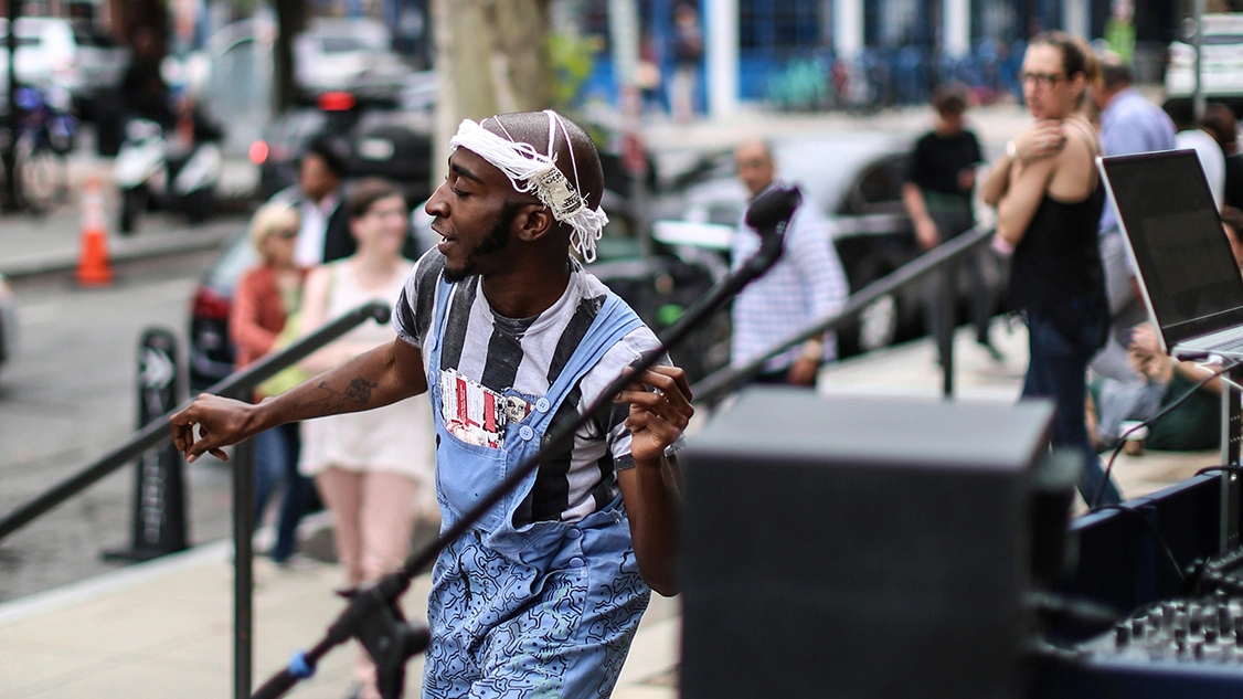 Street performer dancing with mic stand in striped shirt and overalls, surrounded by pedestrians and audio gear