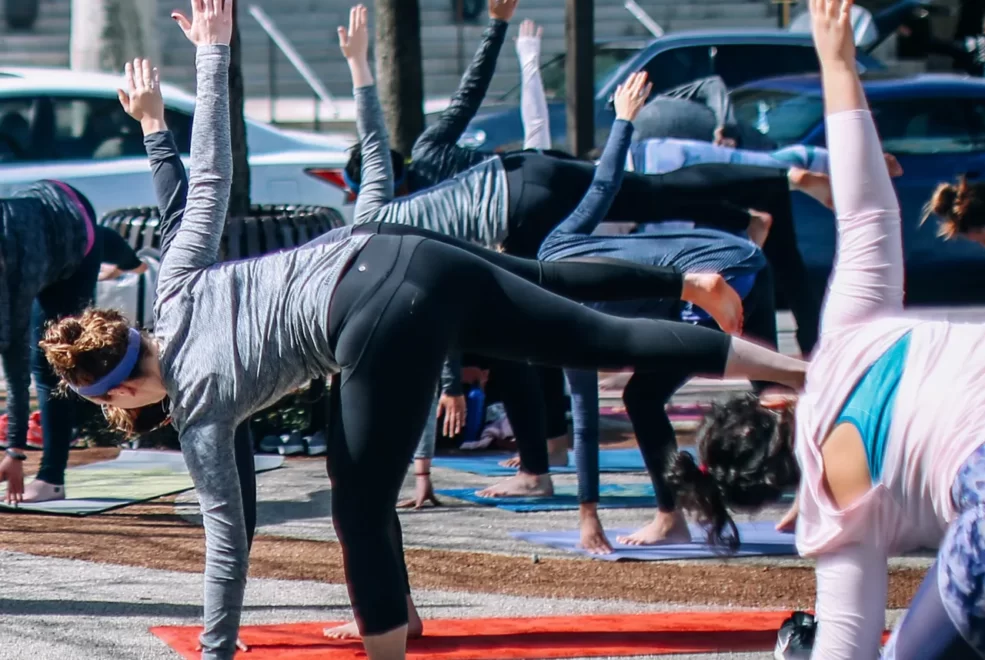 Outdoor yoga class in Half Moon Pose on pavement with rows of mats and urban backdrop