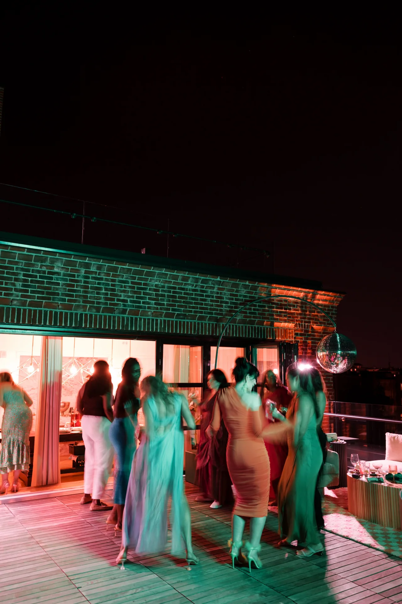 People dancing under disco ball on rooftop terrace with colored lights and warm indoor glow