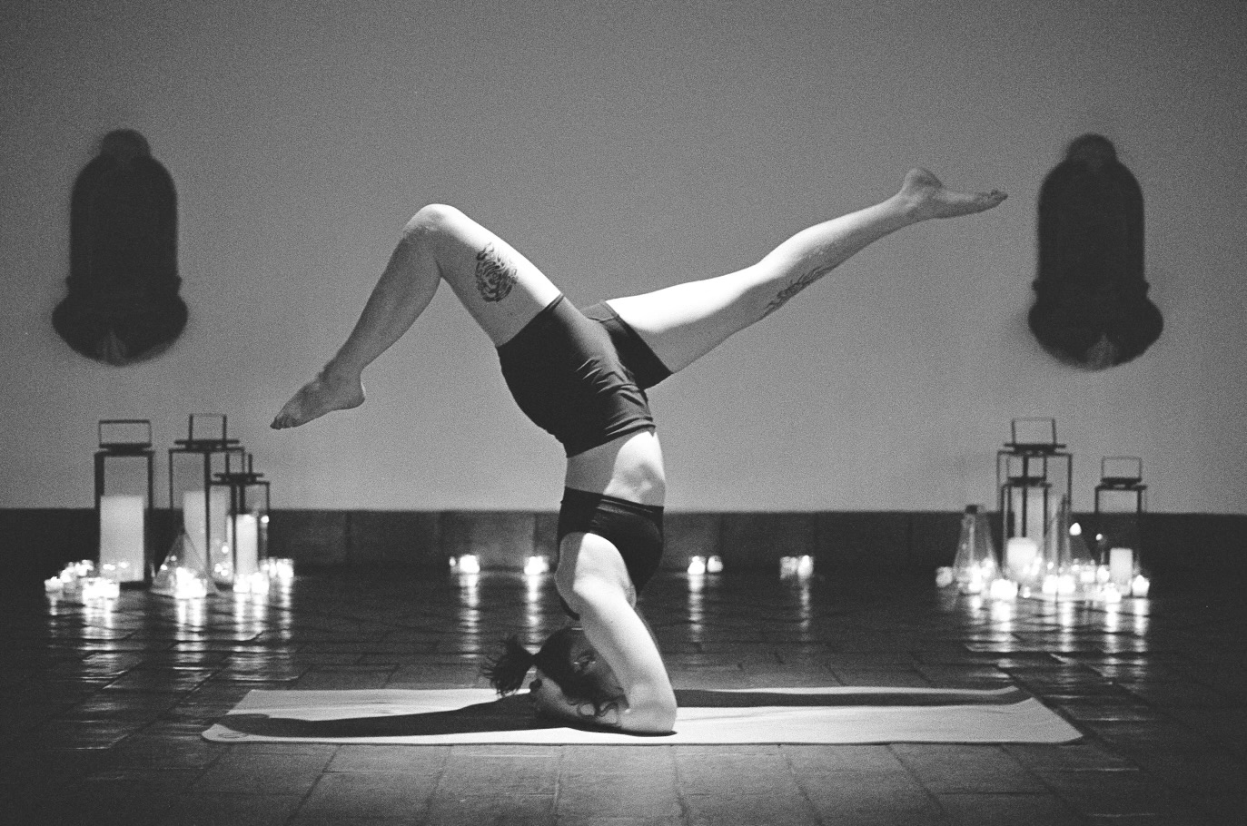 Black and white photo of person balancing in headstand yoga pose with candles in a dimly lit room.