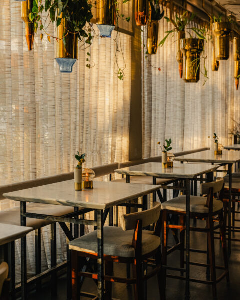 A sunlit restaurant interior with marble tables, high-top chairs, and hanging gold planters against sheer curtains.
