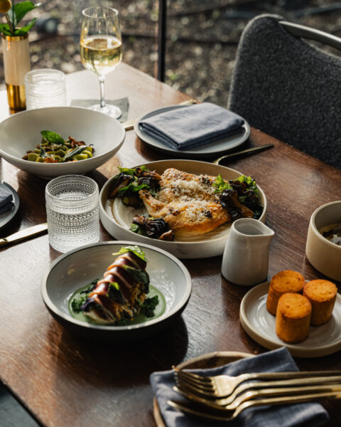 A spread of gourmet dishes, including chicken and pasta, on a wooden restaurant table with white wine.