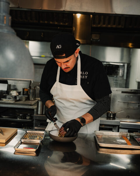 A chef wearing a black cap, apron, and gloves carefully plates a dish on a stainless steel counter in a professional kitchen.