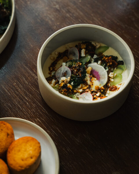 A ceramic bowl filled with a creamy dish topped with broccoli, sliced radishes, nuts, and chili oil on a wooden table.