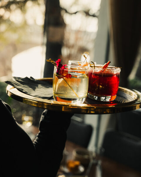 A server carrying a gold tray with three colorful, garnished cocktails in lowball glasses.