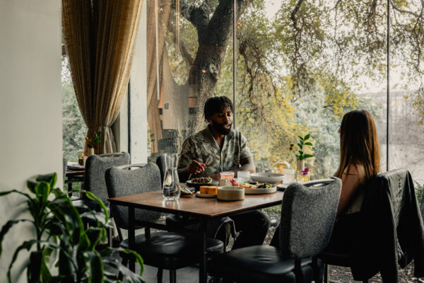 A couple dines at a restaurant table next to a large glass window overlooking trees.