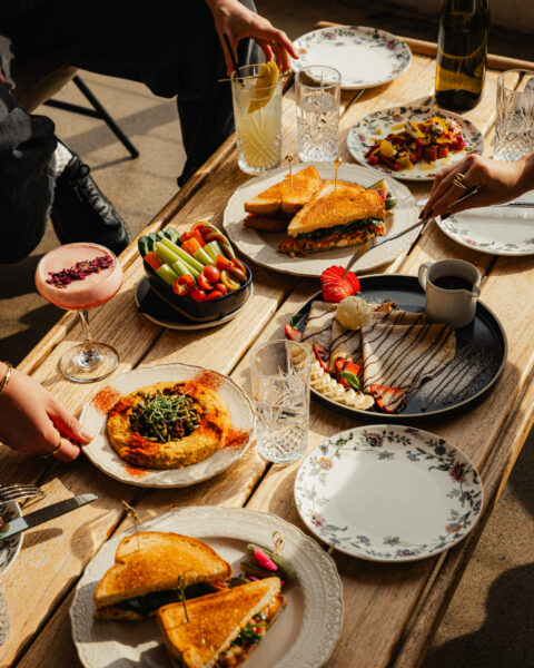 A wooden table spread with brunch dishes including sandwiches, crepes, hummus, a crudité bowl, and cocktails.