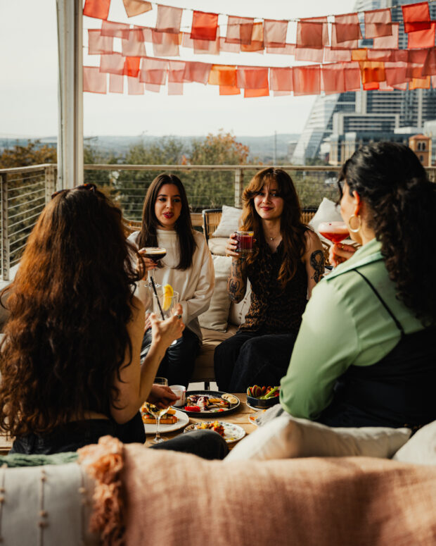 Four friends enjoying cocktails and food on an outdoor rooftop patio with a city view in the background.