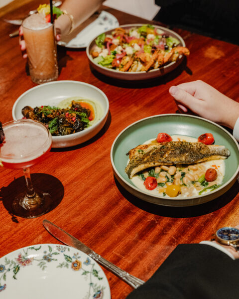 A variety of plated dishes, including fish, salad, and cocktails, served on a wooden dinner table.