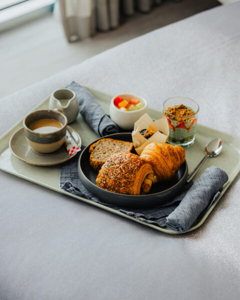 A breakfast tray on a bed featuring croissants, pastries, a cup of coffee, fresh fruit, and a yogurt parfait.