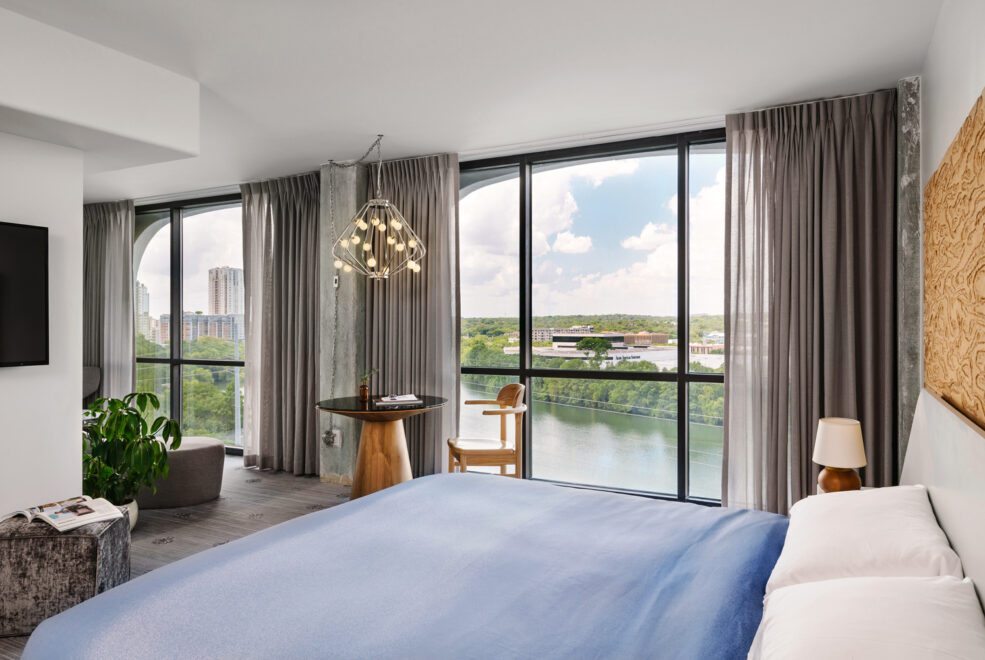 A hotel room features a bed and two floor-to-ceiling windows overlooking a lake