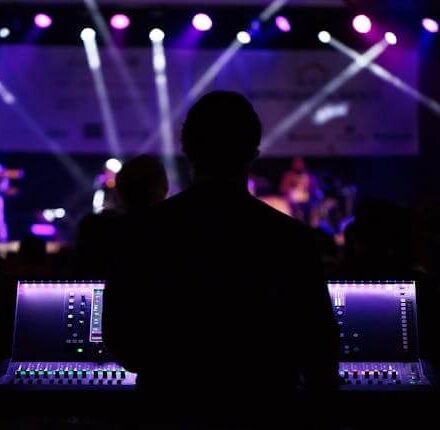 Silhouette of a sound technician working at a live concert, with stage lights and performers in the background.
