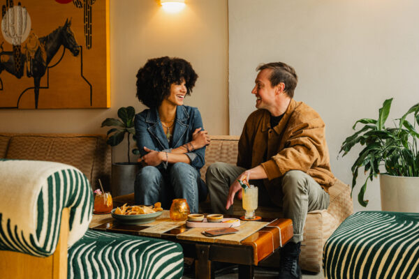 Smiling man and woman sitting on a couch, talking over cocktails and chips in a stylish lounge.
