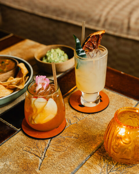 Two garnishing cocktails served on a tiled table next to bowls of tortilla chips and guacamole.