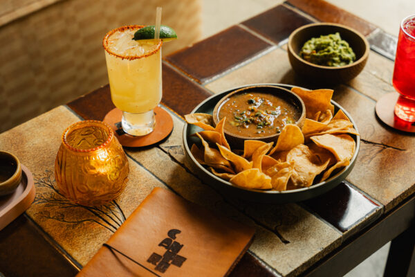 A bowl of tortilla chips and salsa served on a tiled table with a spiced-rim margarita and guacamole.