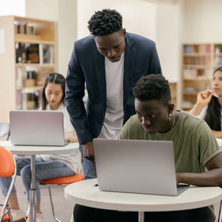 A teacher assists a student on a laptop in a collaborative study space with other focused students nearby