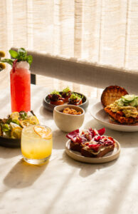 Sunlit table with colorful cocktails and assorted small plates including tacos, salad, and dips.