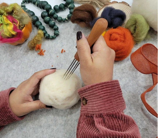 Hands needle-felting white wool on gray mat surrounded by colorful roving, beads, and crafting tools