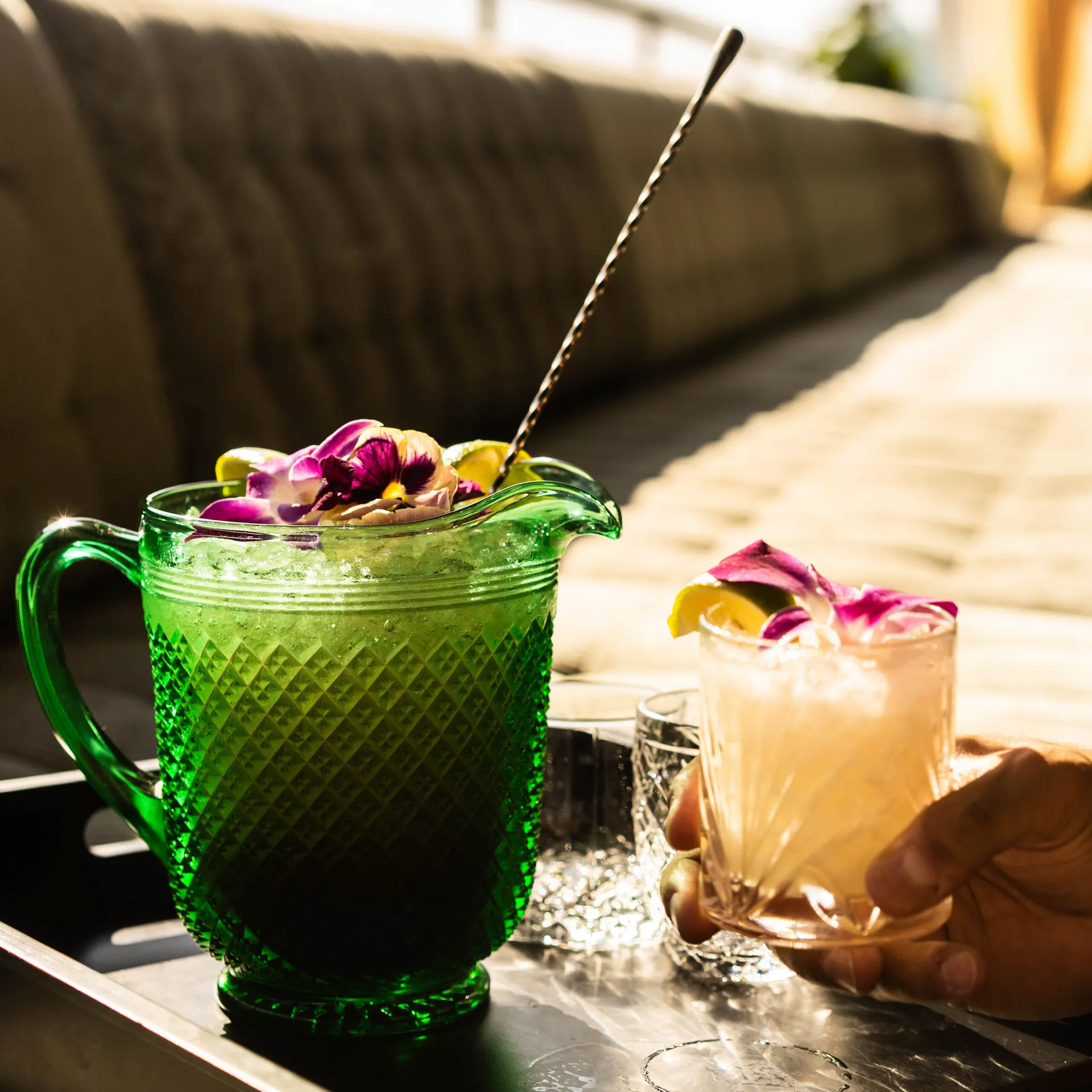 Green pitcher with floral garnish beside hand holding pink drink in sunlit lounge with vintage glassware