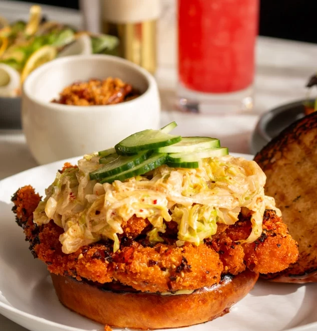 Open fried chicken sandwich with slaw and cucumber beside red drink and colorful side dishes
