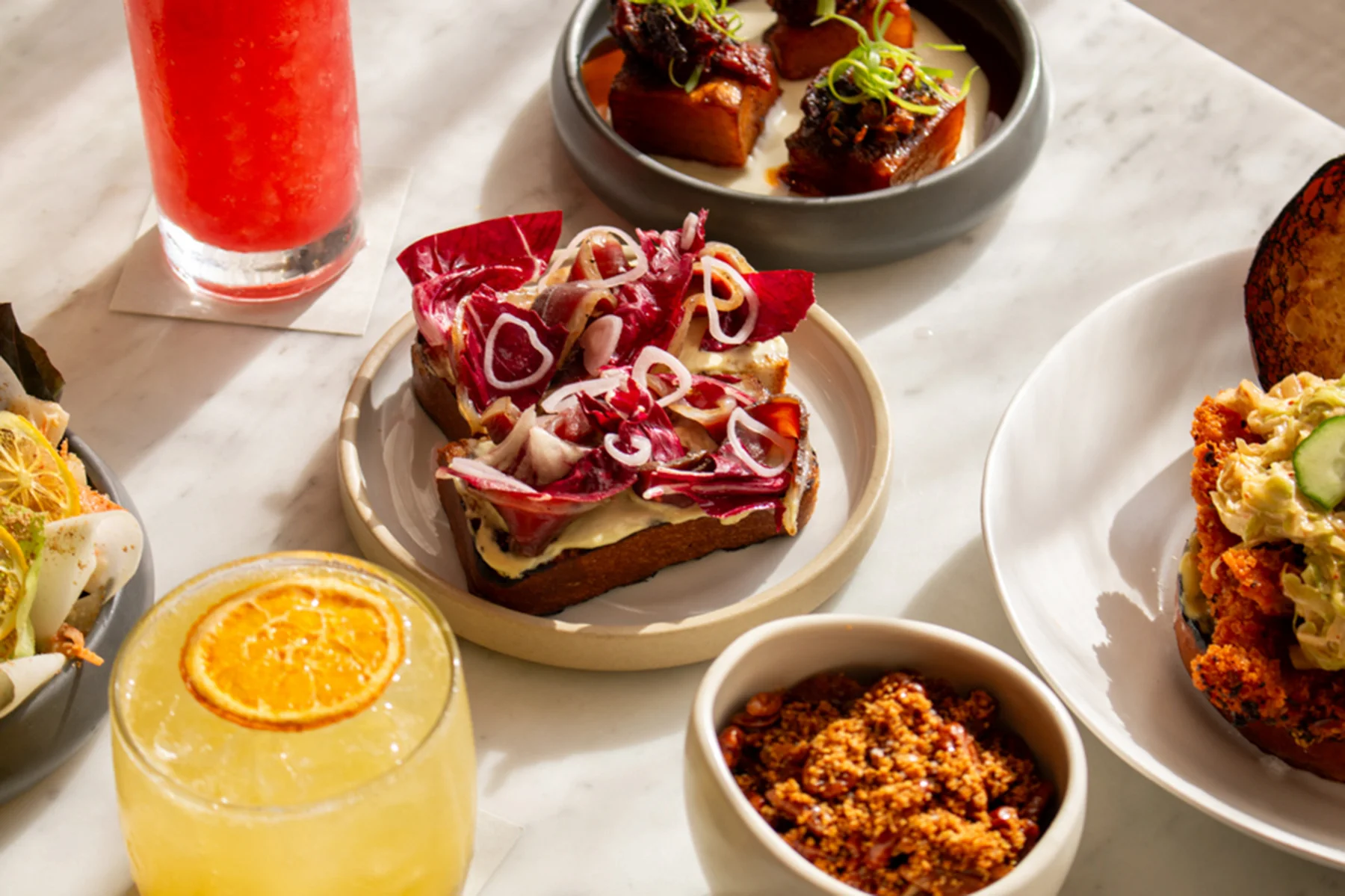 Assorted dishes and drinks on marble table, featuring colorful toast, glazed meat, and fried chicken