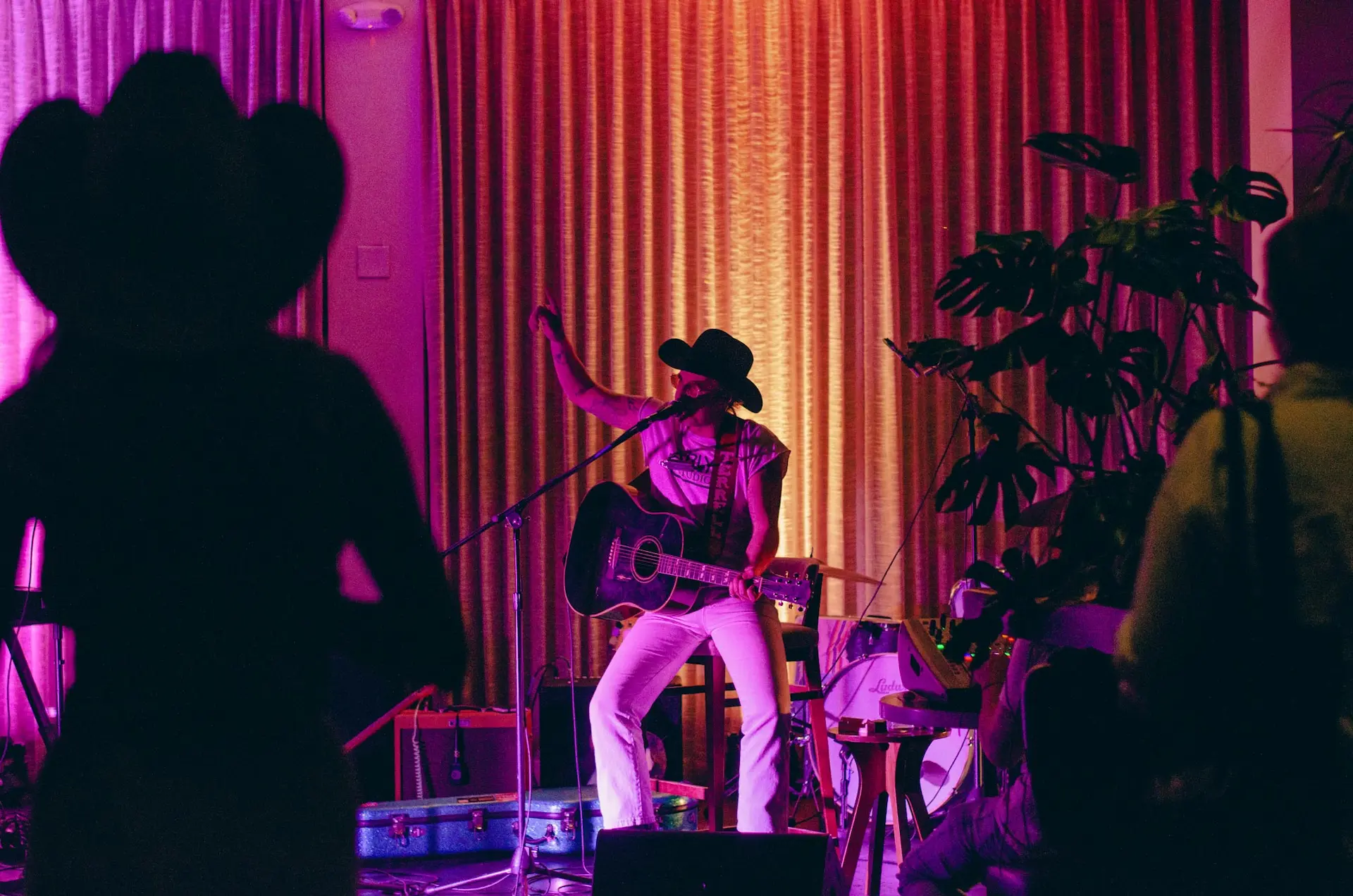 A man plays a guitar while seated in front of a curtain with orange and pink spotlights