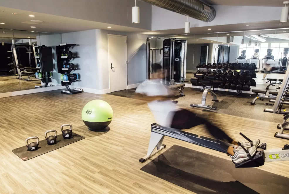 Person rowing in modern gym with kettlebells, green exercise ball, and mirrored weight racks in motion blur