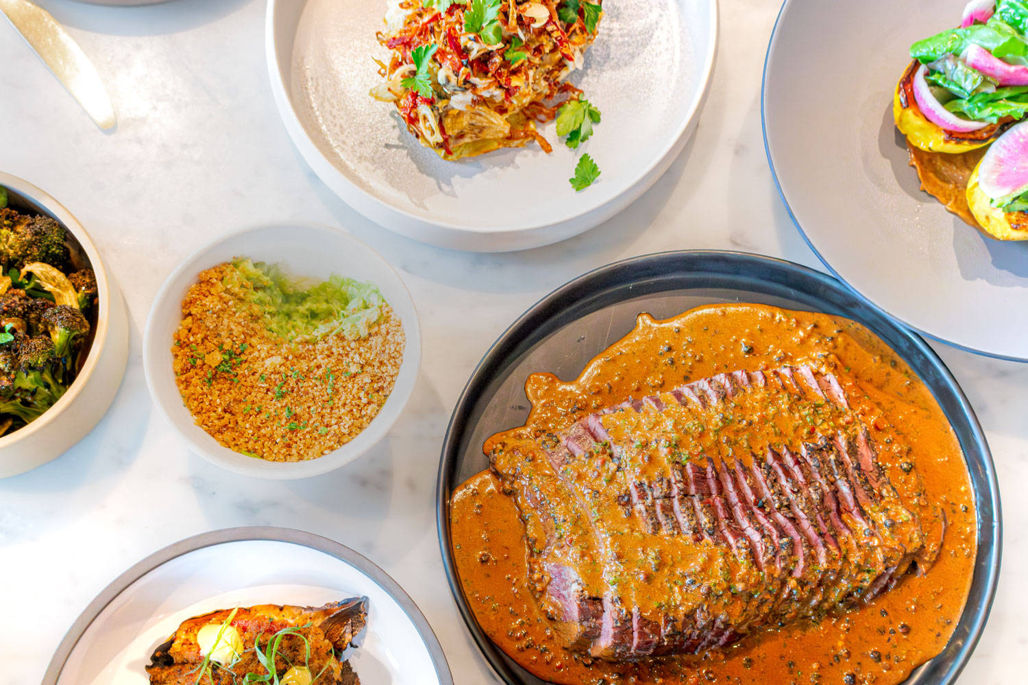 Sliced steak with pepper sauce surrounded by colorful side dishes on marble table