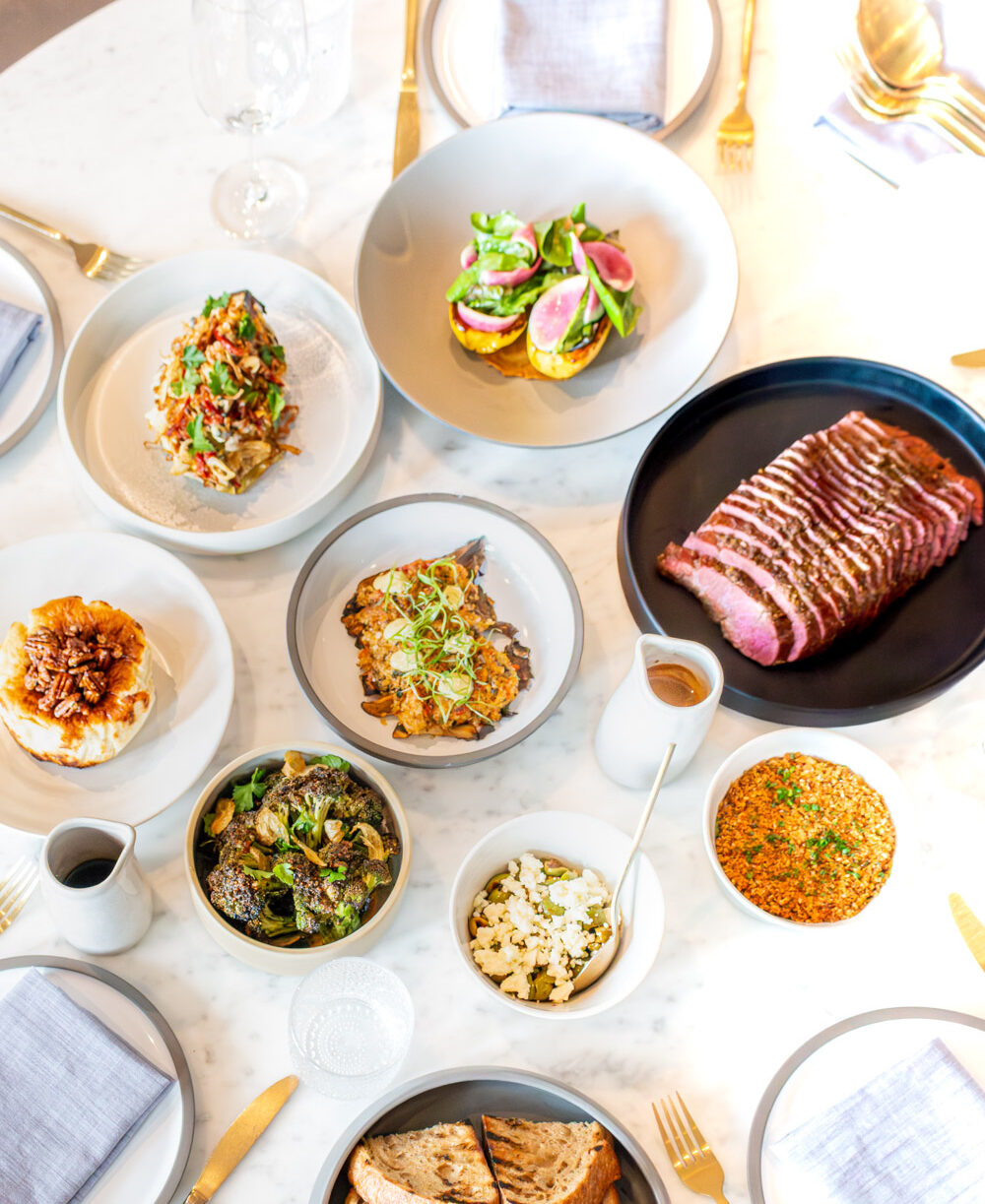 Sliced steak with pepper sauce surrounded by colorful side dishes on marble table.