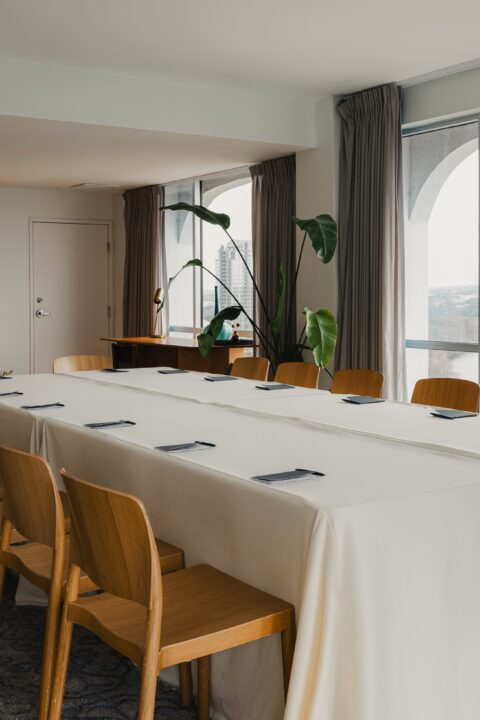 Modern meeting room with long table, wooden chairs, large windows overlooking a river, and a kitchenette in the back.