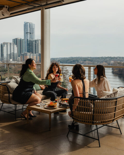 Four women toasting cocktails on a rooftop terrace with city and river views.